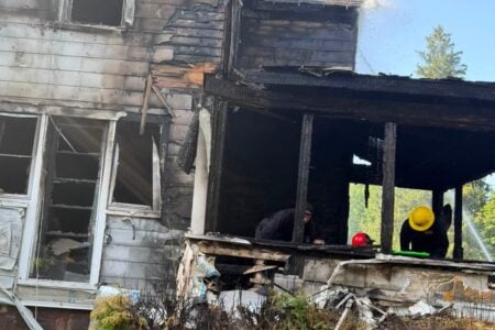 A firefighter sprays water on a severely charred house, with another person visible inside the damaged porch structure.
