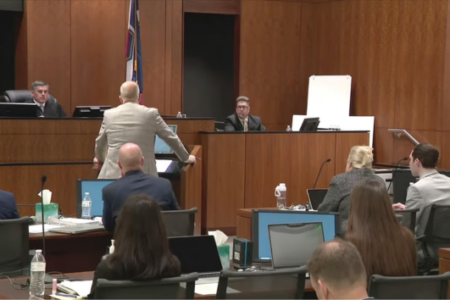 An attorney in a light suit addresses a judge and jury in a wood-paneled courtroom.