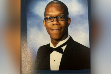 Young Black man wearing glasses and a tuxedo smiles in a formal portrait against a blue and white background.