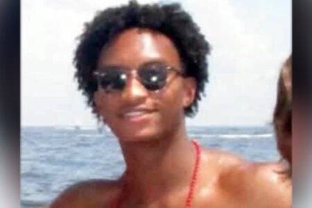 Smiling person with curly hair and sunglasses against a water background with a boat in the distance.