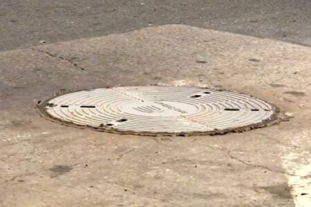 A round Syracuse manhole cover with concentric circles is embedded in cracked asphalt and concrete pavement.