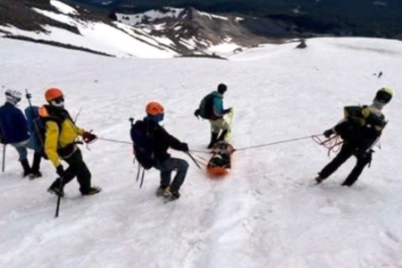 A mountain team in climbing gear pulls an orange rescue sled down a snow-covered slope.