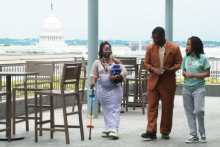 A man in a suit, a woman with a rainbow cane, and a woman in a "SOUL" t-shirt walk on a rooftop overlooking the Capitol.