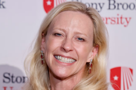 A blonde woman with blue eyes smiles, wearing red and silver earrings, in front of a Stony Brook University logo.