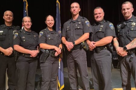 Six police officers in dark uniforms stand on a stage with flags behind them.