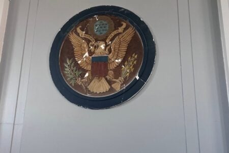 Weathered Great Seal of the U.S. emblem on a gray wall with decorative trim.