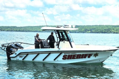 Two Ontario County Sheriff's deputies patrol a lake in their boat, with forested hills in the background under a blue sky.