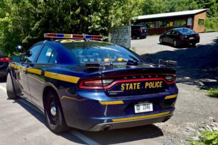 New York State Police car parked on pavement near a small building and green trees.