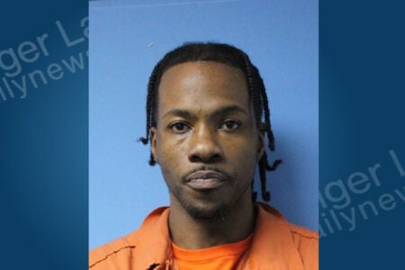 Mugshot of an African American man with braided hair and a goatee, wearing an orange uniform against a blue wall.