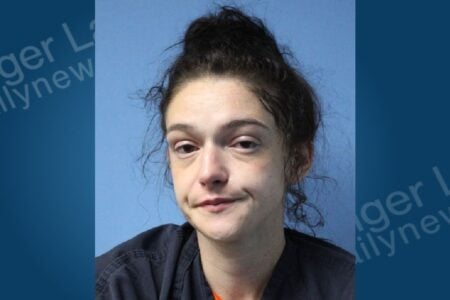Woman with messy dark hair in a bun, slight smirk, wearing a dark uniform, looks ahead in a mugshot on a blue background.