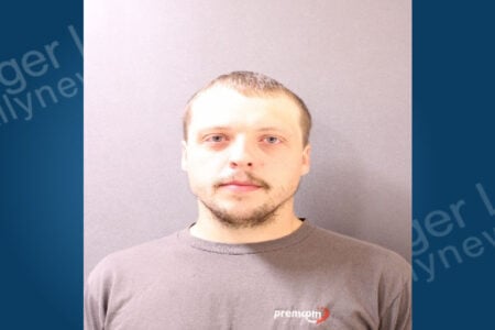 Mugshot of a man with light hair, blue eyes, and a beard, wearing a grey t-shirt, against a light background.