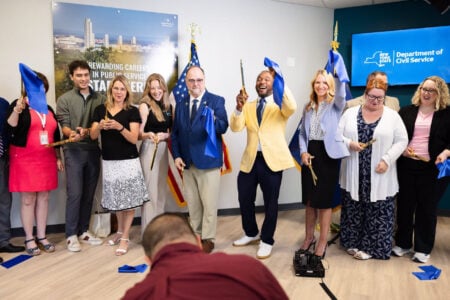 Group cuts a blue ribbon with large scissors at a New York State Department of Civil Service office opening.