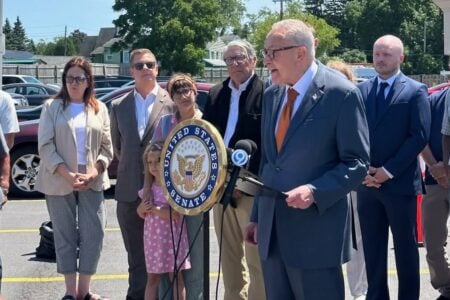 Senator Chuck Schumer speaks at an outdoor press conference with a Senate seal podium, joined by community members.