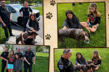 Two girls pet a playful brown and white speckled dog as police officers stand by on a sunny day outdoors.