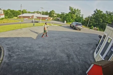 Man in shorts and t-shirt walks across an asphalt parking lot, holding a phone, with a street and buildings behind him.