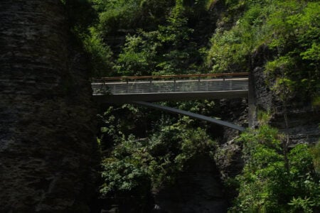 Modern metal and wood pedestrian bridge spanning a deep, rocky gorge with lush trees.