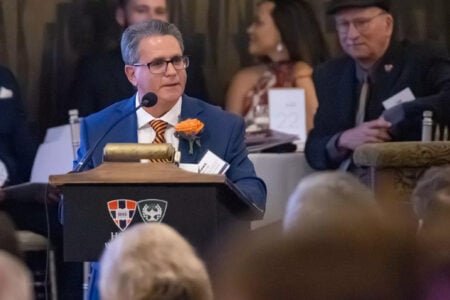 A man in a blue suit and orange tie speaks at a HWS podium to an audience at a formal indoor event.