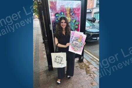 Smiling woman in black stands on a brick sidewalk, holding colorful art prints and a tote bag, by an outdoor art installation.