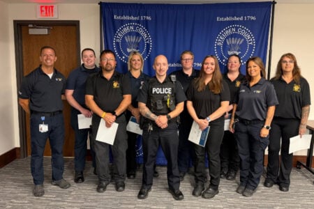 Eleven Steuben County emergency responders and one police officer pose with certificates in front of a Steuben County banner.
