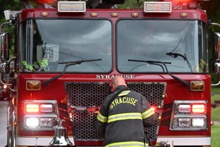 Syracuse firefighter works at the front of a red Syracuse fire truck with its emergency lights flashing.