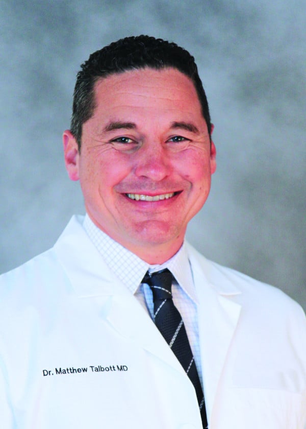 Dr. Matthew Talbott MD smiles in a white lab coat, his name embroidered, against a grey background.