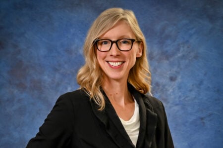Blonde woman in glasses smiling at the camera, wearing a black blazer against a blue textured background.