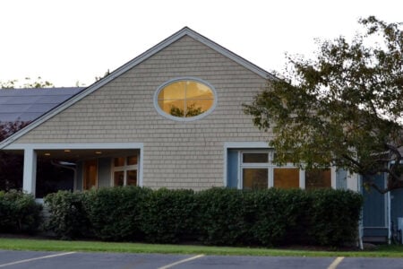 Tan shingled building with a prominent oval window, covered entrance, and solar panels on its gabled roof.