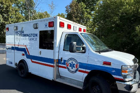 White Victor Farmington Ambulance with blue and red stripes parked outdoors against green trees.