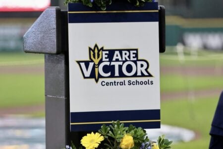 A podium displays the "WE ARE VICTOR Central Schools" sign with a trident logo, set on a baseball field.