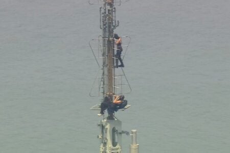 Two individuals climb a tall metal tower, one pausing on a platform, high above calm water.