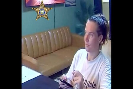 A person in a white 'Angels' shirt holds a phone and pen, looking right while seated indoors next to a tan couch.