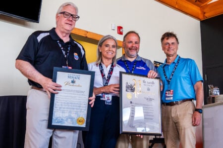 Four people smile while holding New York Senate and Assembly proclamations for NASCAR at Watkins Glen International.