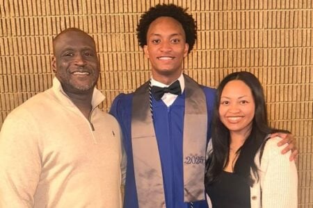 Student in blue graduation gown and '2025' stole smiles with two adults against a textured wall.