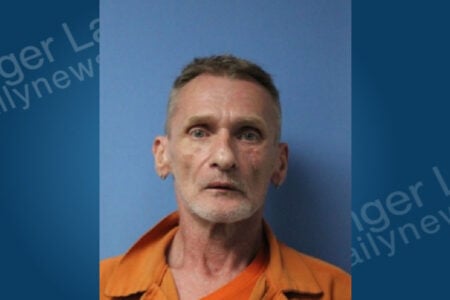Mugshot of a man with short graying hair and a goatee, wearing an orange jumpsuit, against a blue background.