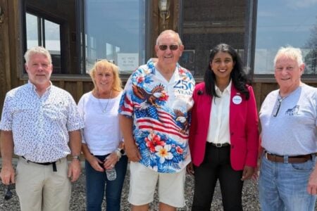 Five individuals, including a woman in a pink blazer wearing a 'Saritha' button, stand outside a wooden building.
