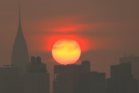 Red-orange sun glows through dense haze behind the NYC skyline, including the Chrysler Building.