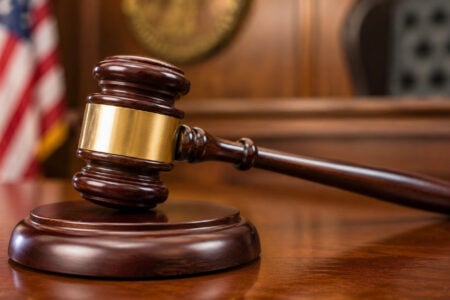 A dark wooden gavel with a brass band rests on its block on a polished table in a courtroom setting.