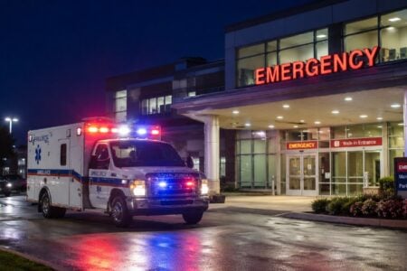 An ambulance with emergency lights pulls up to a hospital emergency room entrance under a red "EMERGENCY" sign at night.