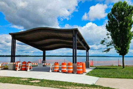 Wavy-roof pavilion blocked by orange barrels and "sidewalk closed" signs, next to a Finger Lake.