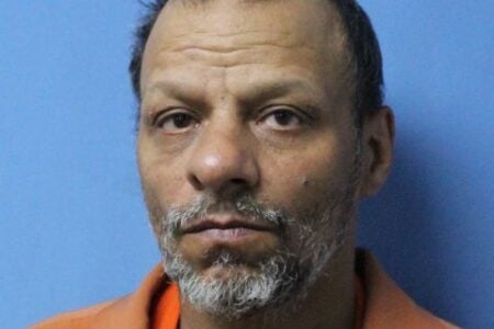 Mugshot of a man with balding dark hair, a grey beard, and an orange jumpsuit, looking directly forward.