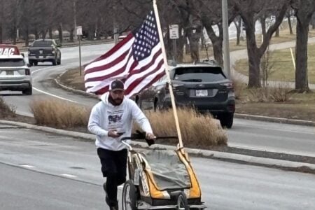 Bearded man in a hoodie runs on a road, pushing a cart with a large American flag attached.