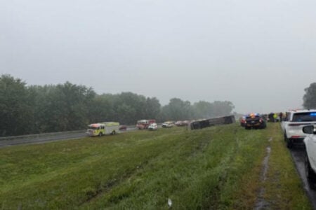 Overturned semi-truck on a rainy highway embankment with fire trucks, police cars, and emergency personnel at the scene.