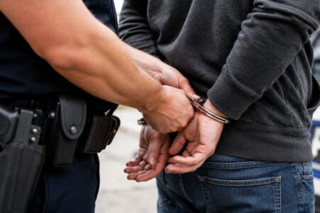Police officer in uniform placing handcuffs on an individual wearing a dark hoodie and jeans, near a police car.