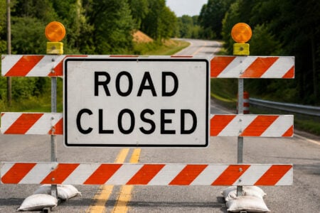 A "ROAD CLOSED" sign on an orange and white striped barrier with flashing lights blocks a tree-lined rural road.