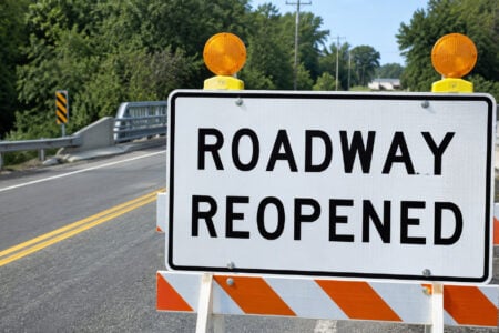 A "ROADWAY REOPENED" sign with two amber lights stands on an asphalt road leading to a bridge.