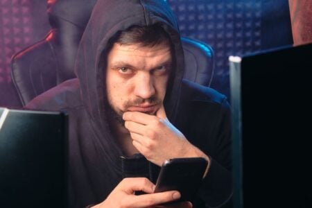Bearded man in dark hoodie looks intently at the viewer, holding a smartphone near multiple screens in a neon-lit room.