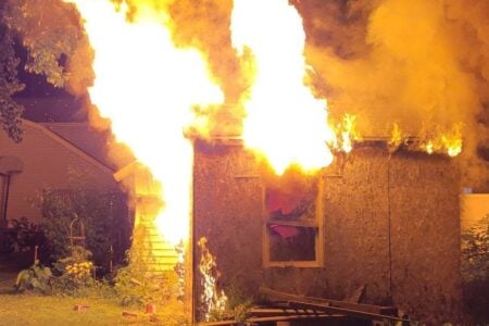 Wooden outbuilding fully engulfed in large, bright flames in a residential backyard.