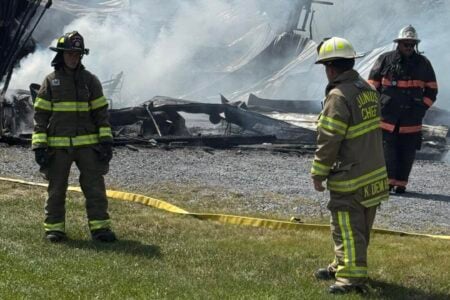 Firefighters, including Junius Chief, stand near a smoking, destroyed building with yellow tape in the foreground.
