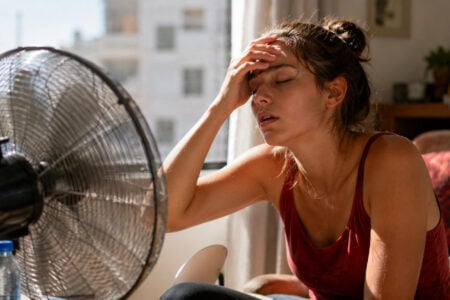 Woman looking hot and wiping her forehead, sitting near a large fan indoors on a sunny day.